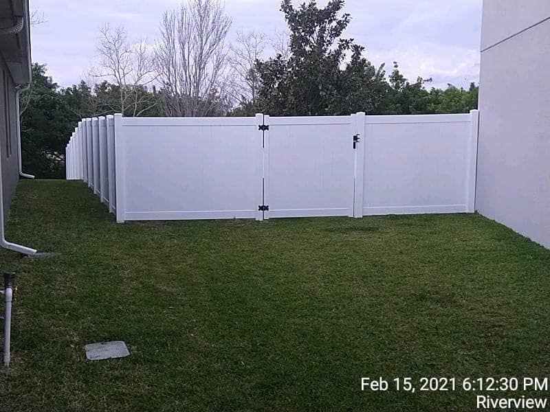 White vinyl fence encloses a grassy backyard with a gate, trees, and house exterior.