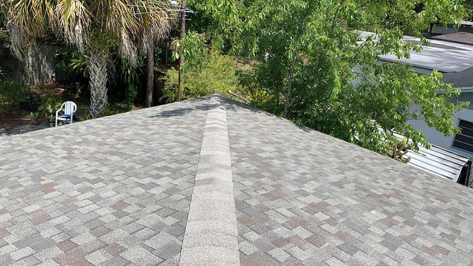 Close-up of a roof with shingles in shades of brown and gray; trees and a chair are visible in the background.