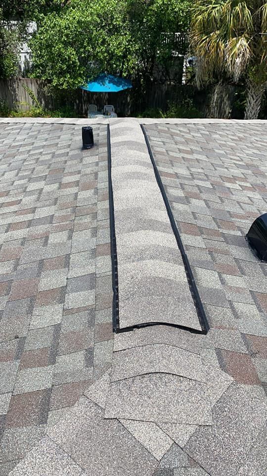 Roof with dark seam and shingles in various shades of gray and brown. Blue umbrella in background.