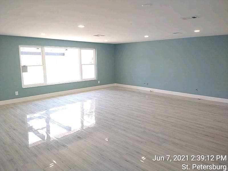 Empty, newly renovated room with light blue walls, white trim, and a window. Reflective, gray wood-look flooring.
