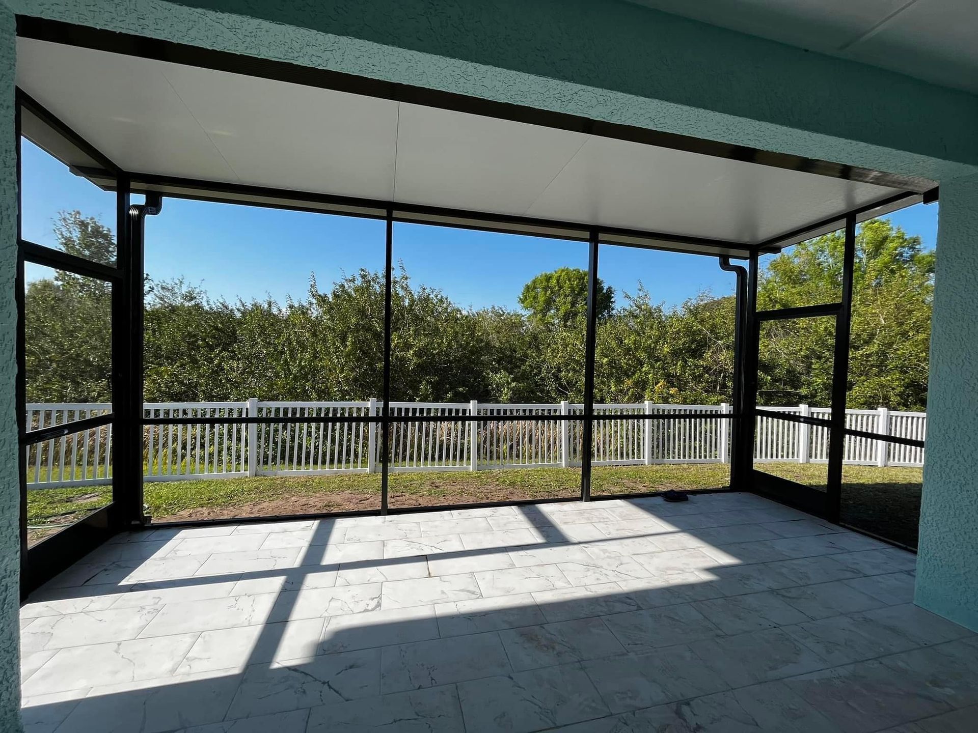 Screened-in patio with black framing and white ceiling. Overlooks a yard with trees and white fence, blue sky.