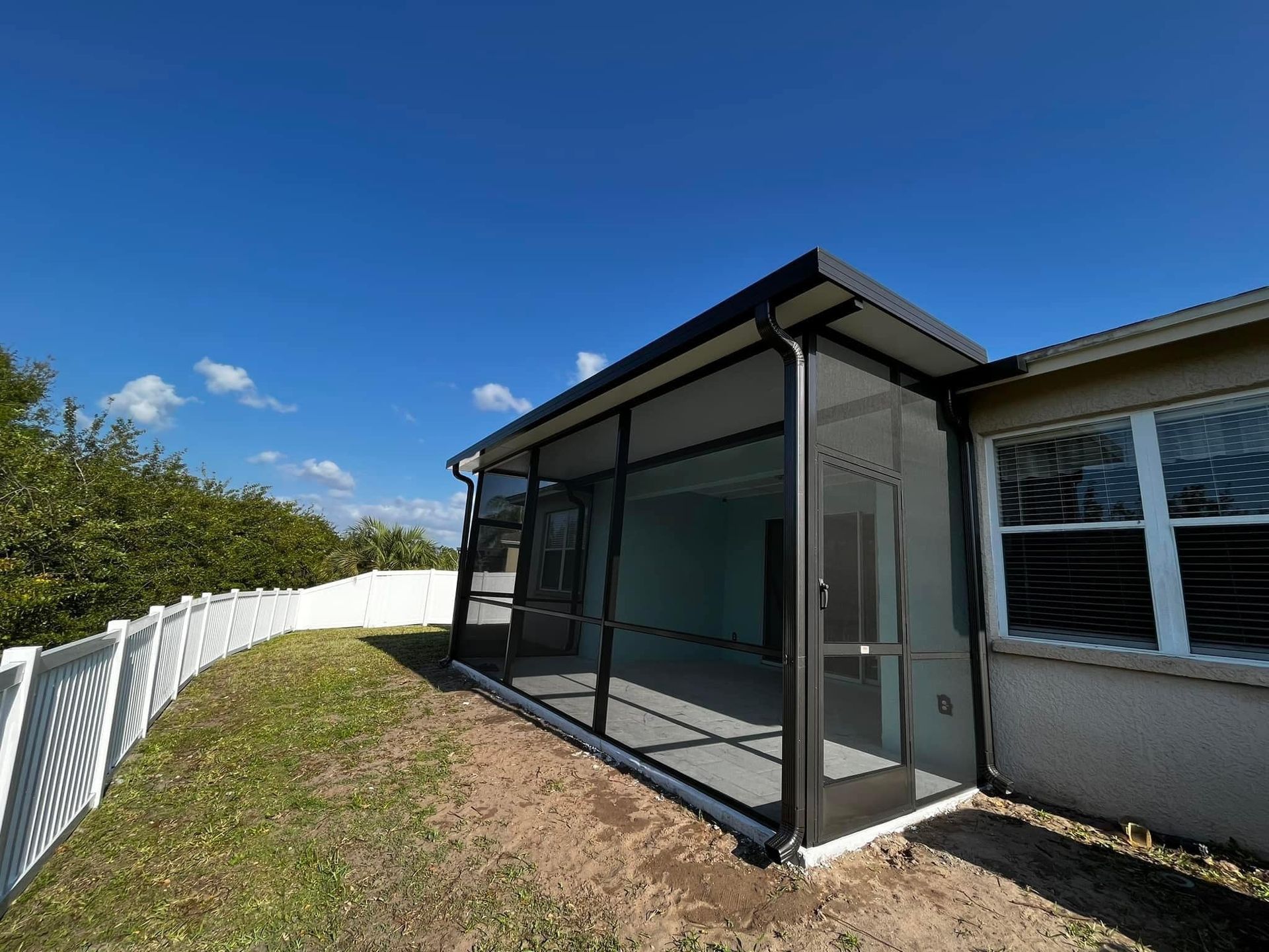 Screened-in porch attached to a house. Black frames with gray floor and blue sky.