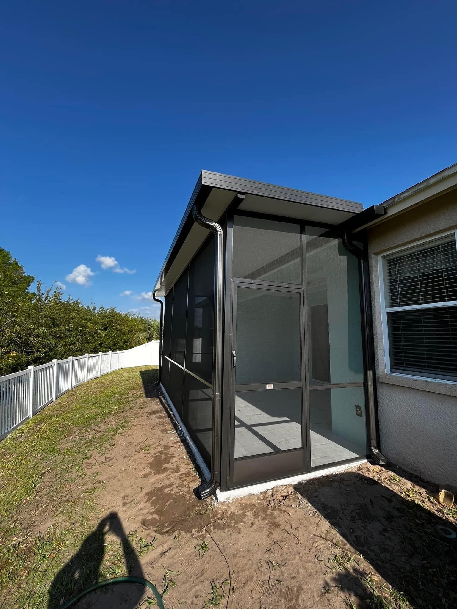 Screened-in patio extension attached to a house with a dark frame, surrounded by a yard and white fence under a blue sky.