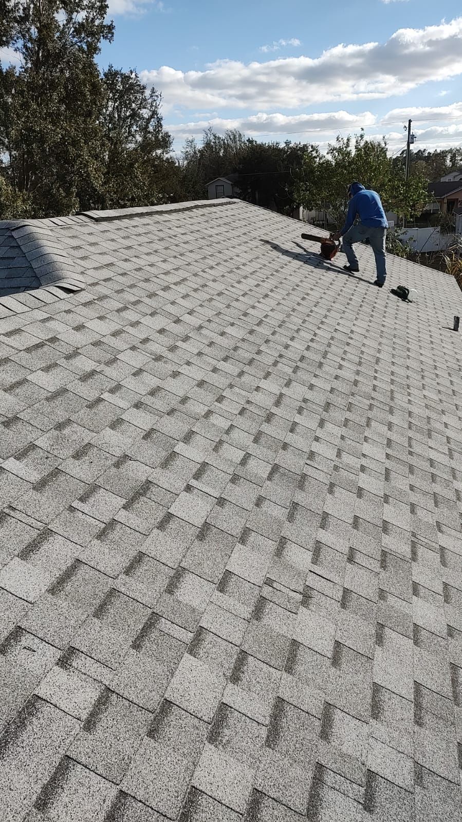 Roofer in blue shirt working on a gray shingled roof under a partly cloudy sky.