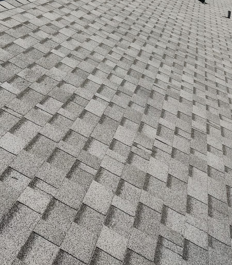 Roofer in blue shirt working on a gray shingled roof under a partly cloudy sky.