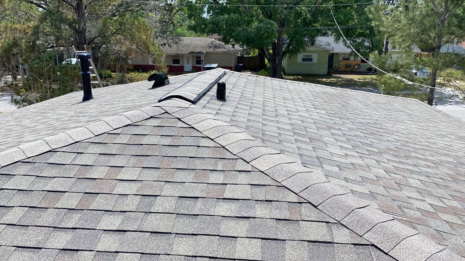 Close-up of a shingled roof, light gray shingles, two black chimneys, and other houses in the background.