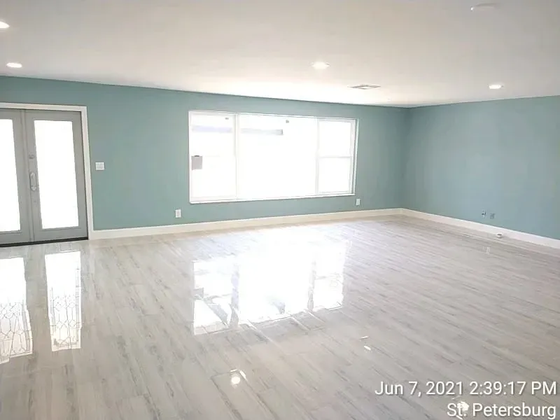 Empty room with light blue walls, white trim, and glossy wood-look floors. Large windows and double doors provide sunlight.