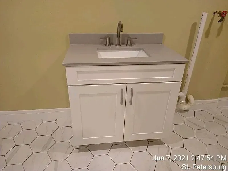 White bathroom vanity with gray countertop and silver faucet, on white hexagon tile floor.