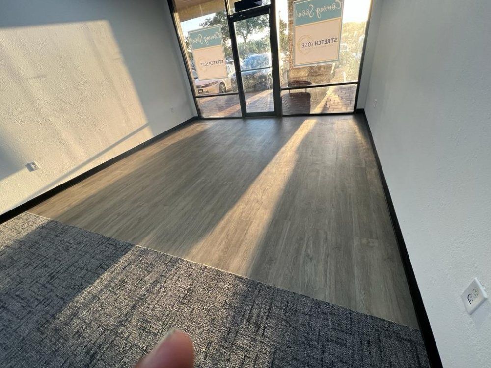 Empty room with wood-look flooring and a grey carpet, sunlight streaming in from the large windows.