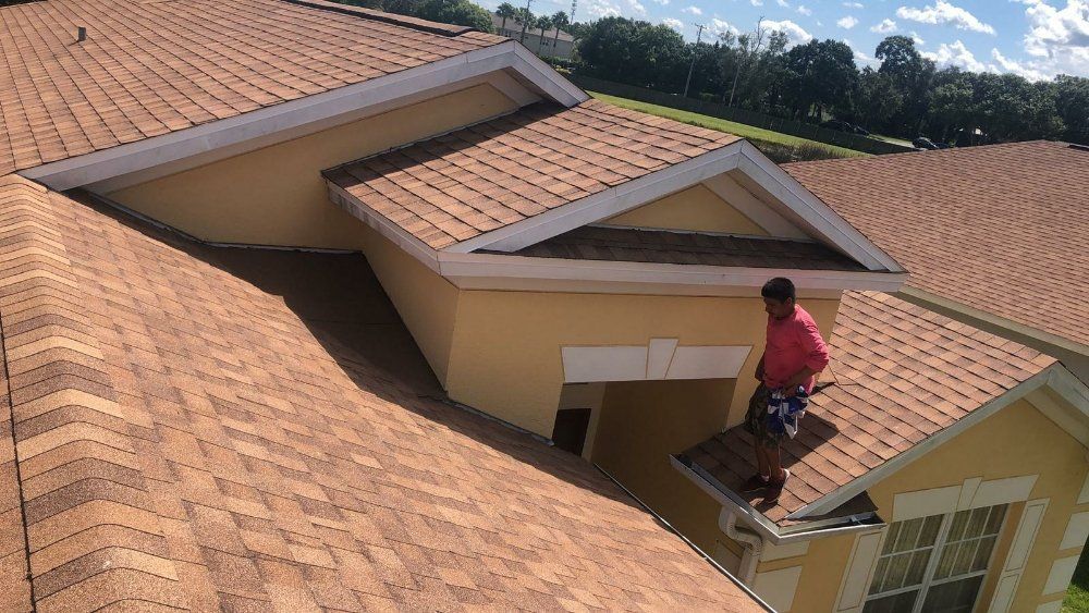 Person on a brown shingle roof, working on a house with a tan exterior. Blue sky in background.