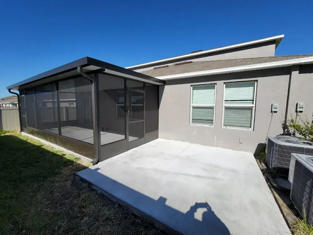 Screened patio next to a gray house, with concrete patio and clear blue sky.