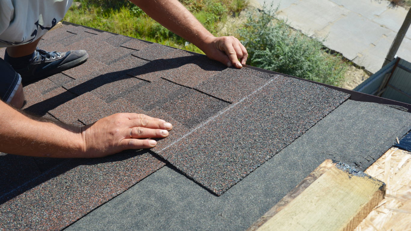 A person installing brown asphalt shingles on a roof over black felt underlayment.