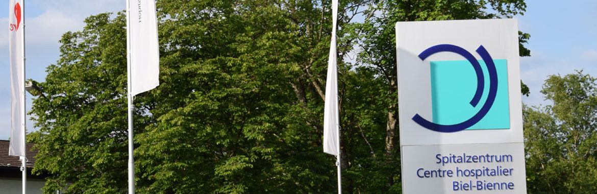 Schild für ein Krankenhaus mit einem blaugrünen D in einem blauen Wirbel vor einem Baum.