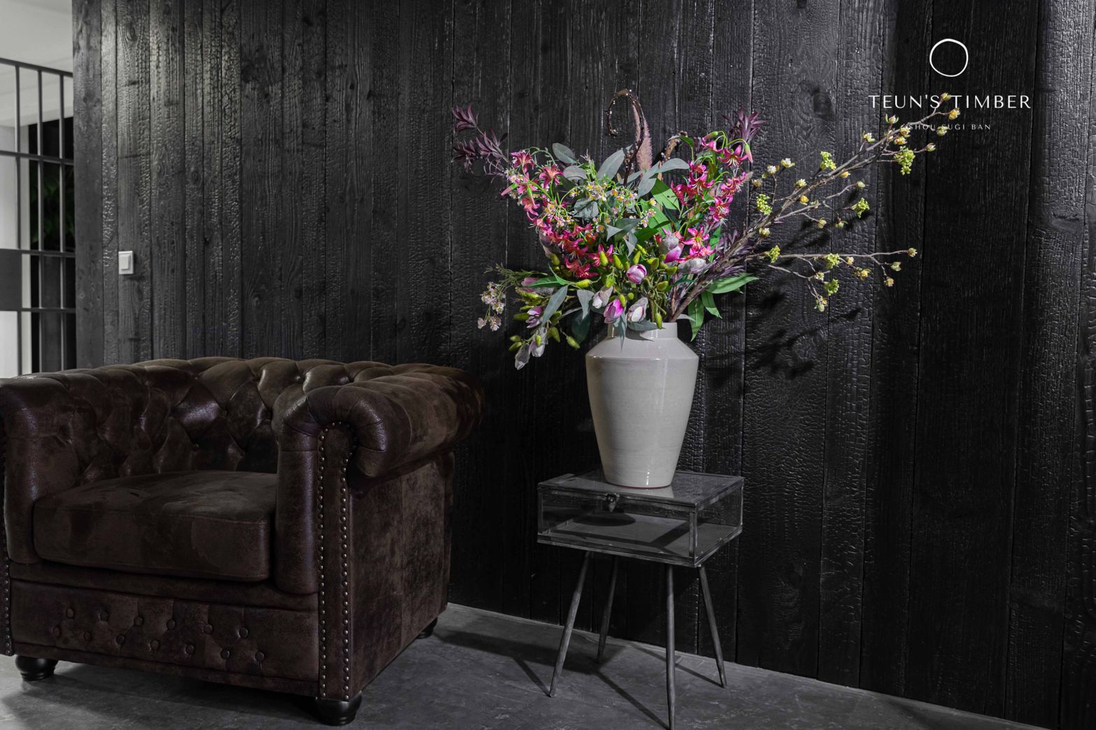 Een vaas met bloemen staat op een tafel naast een stoel voor een shou sugi ban muur