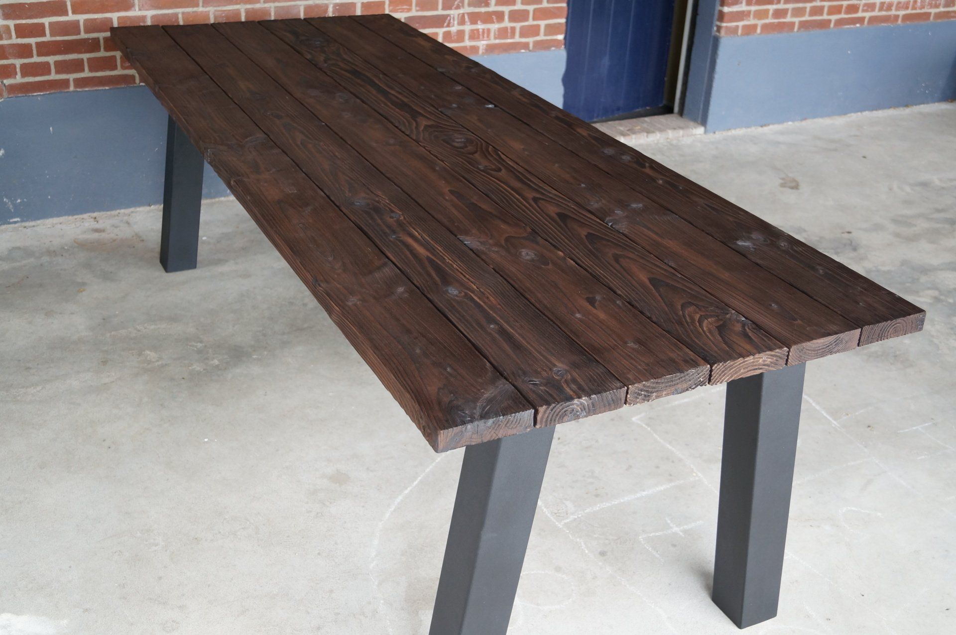 Een houten tafel met zwarte poten staat voor een bakstenen gebouw