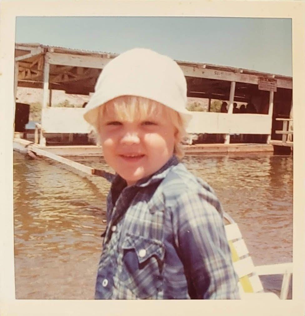 A young boy wearing a plaid shirt and a white hat stands in front of a body of water