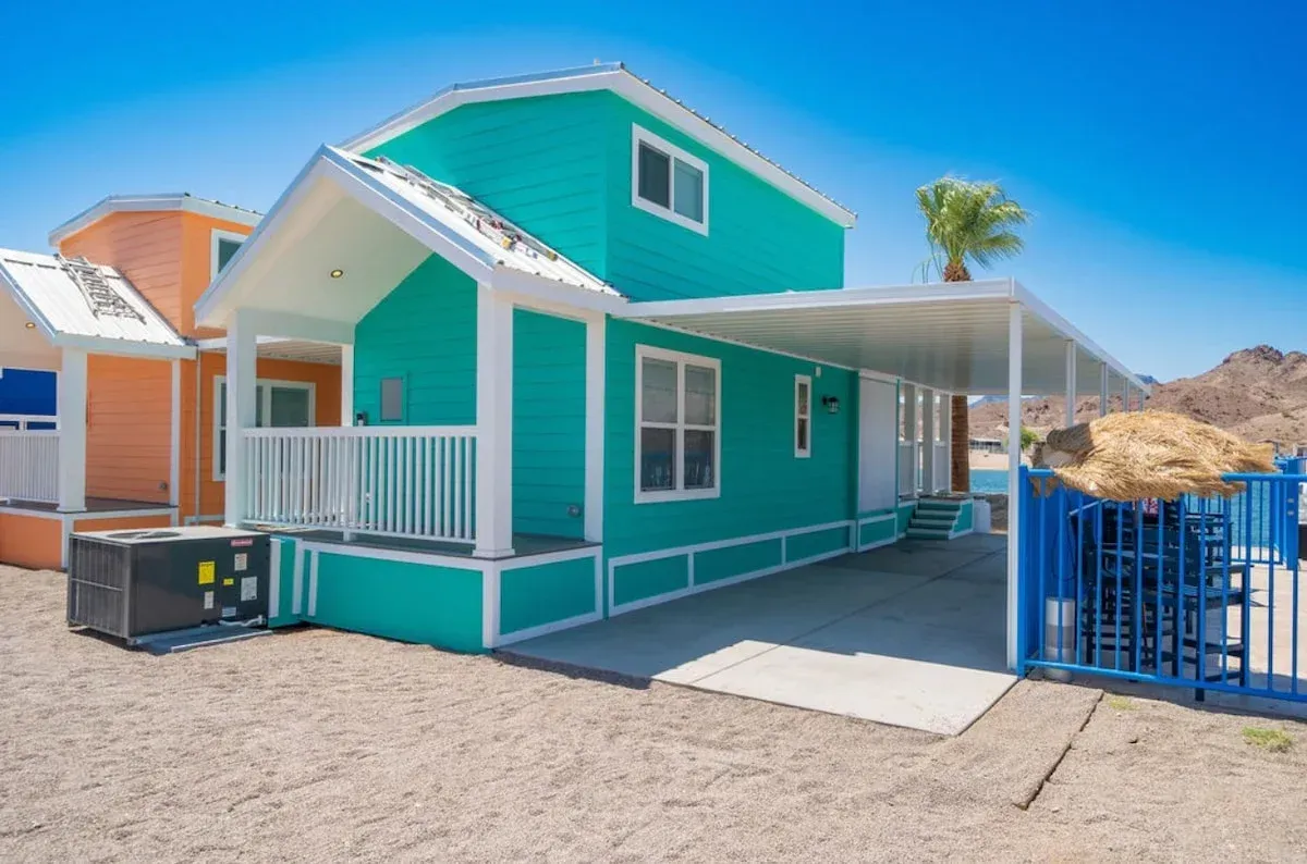 A green house with a white porch is sitting on top of a dirt field.