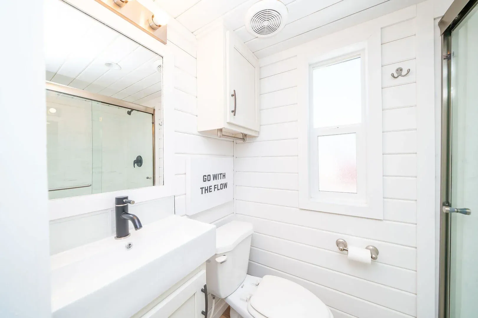 A bathroom with a toilet , sink , mirror and window.