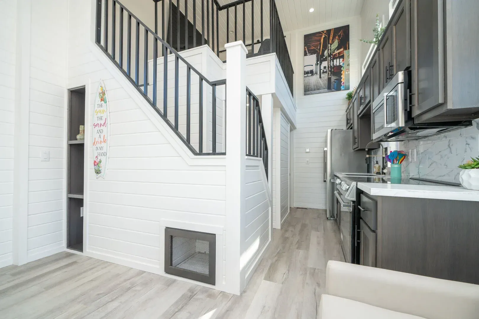 A living room with stairs leading up to the second floor and a kitchen with stainless steel appliances.