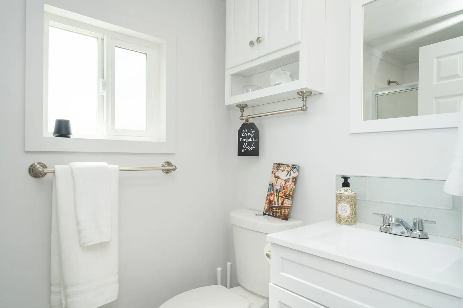 A bathroom with a toilet , sink , mirror and window.
