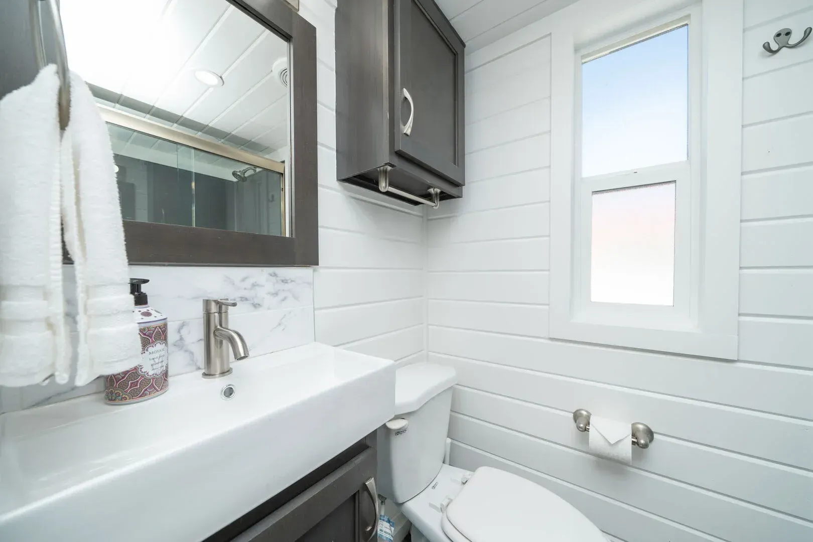 A bathroom with a toilet , sink , mirror and window.