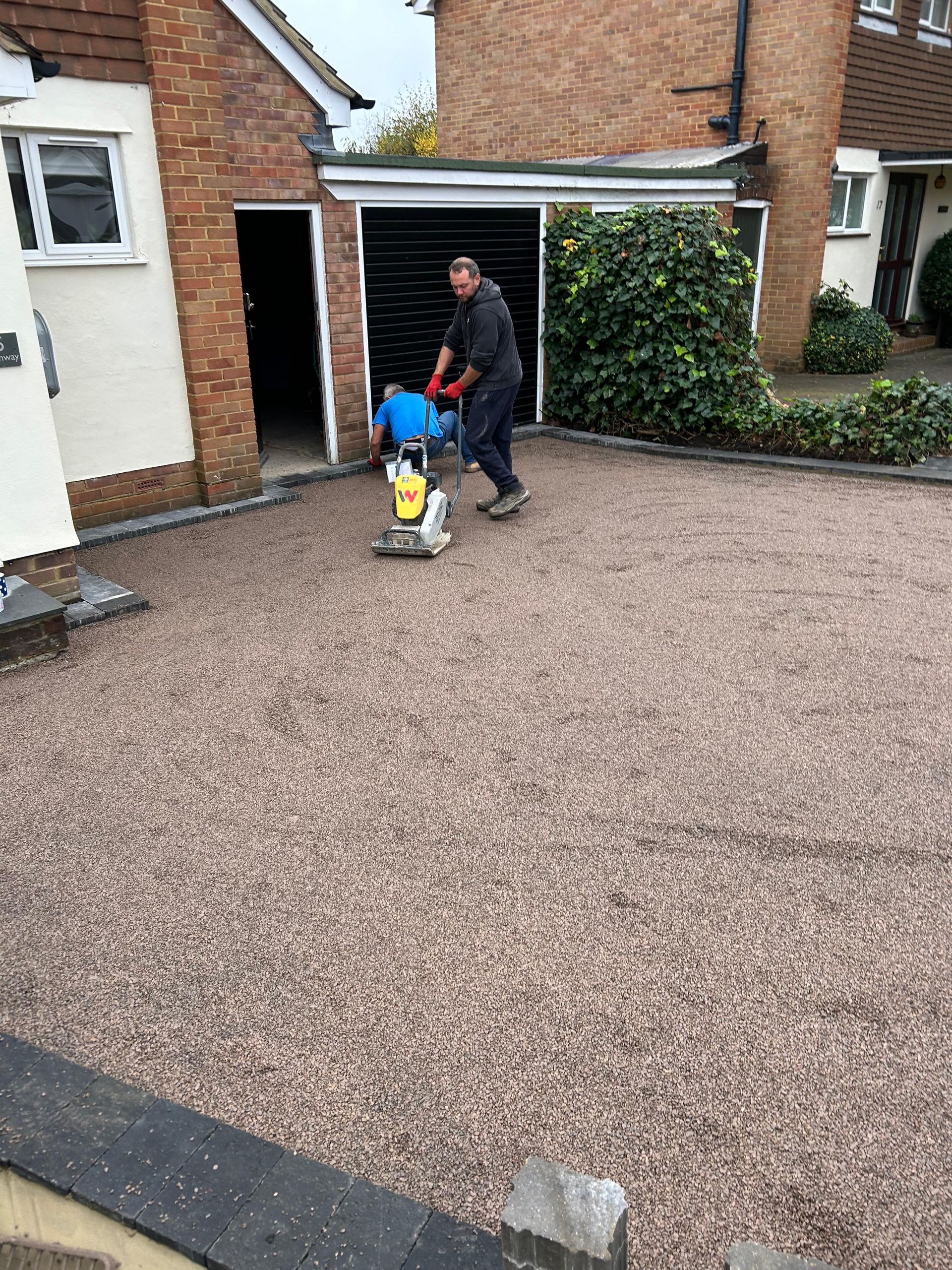 Two men are working on a gravel driveway in front of a house.