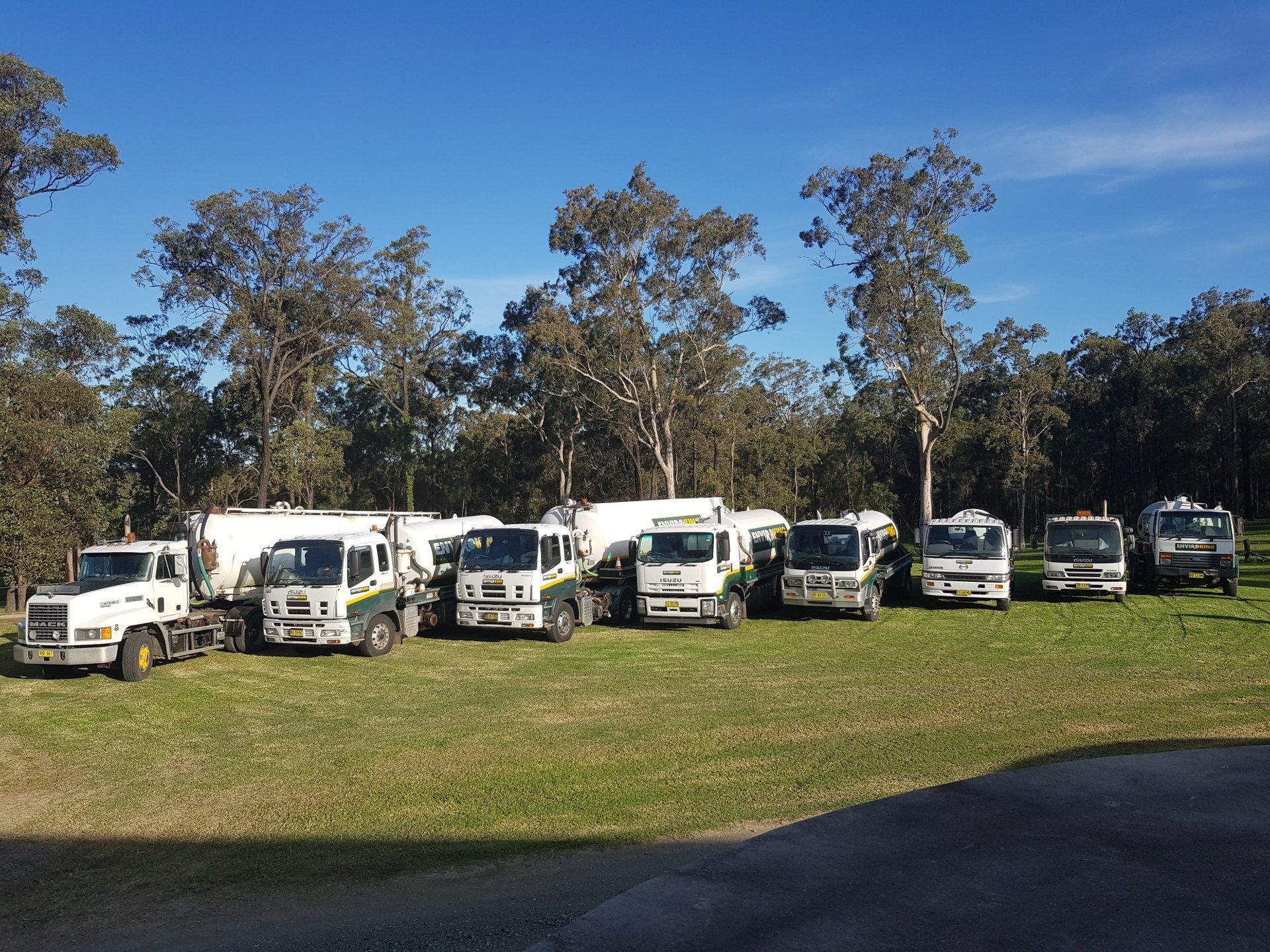 A line of seven white tanker trucks parked on a grassy field with trees in the background.