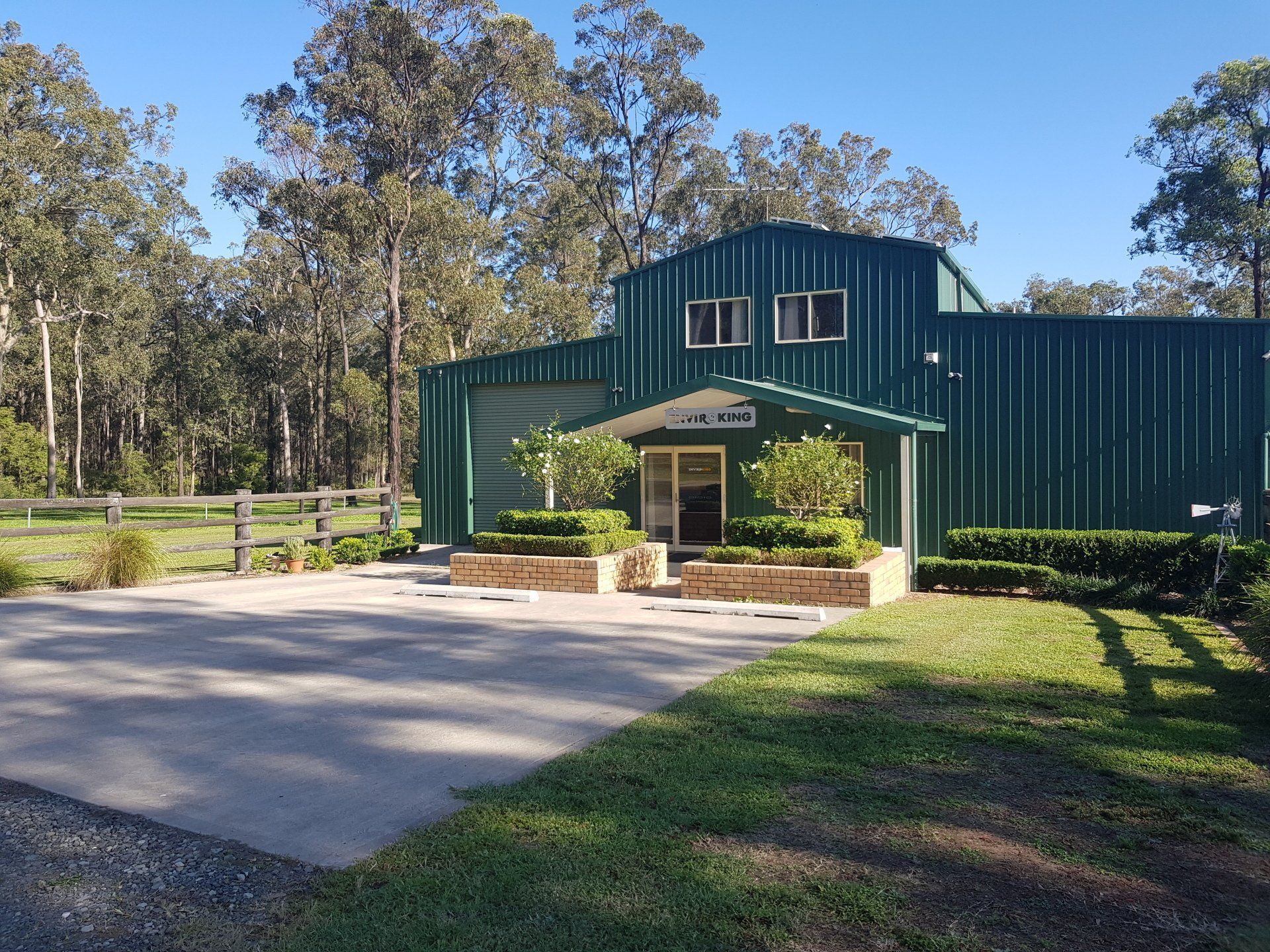 Green metal building with small entryway and landscaped plants. Asphalt driveway and grassy area. Trees in background.