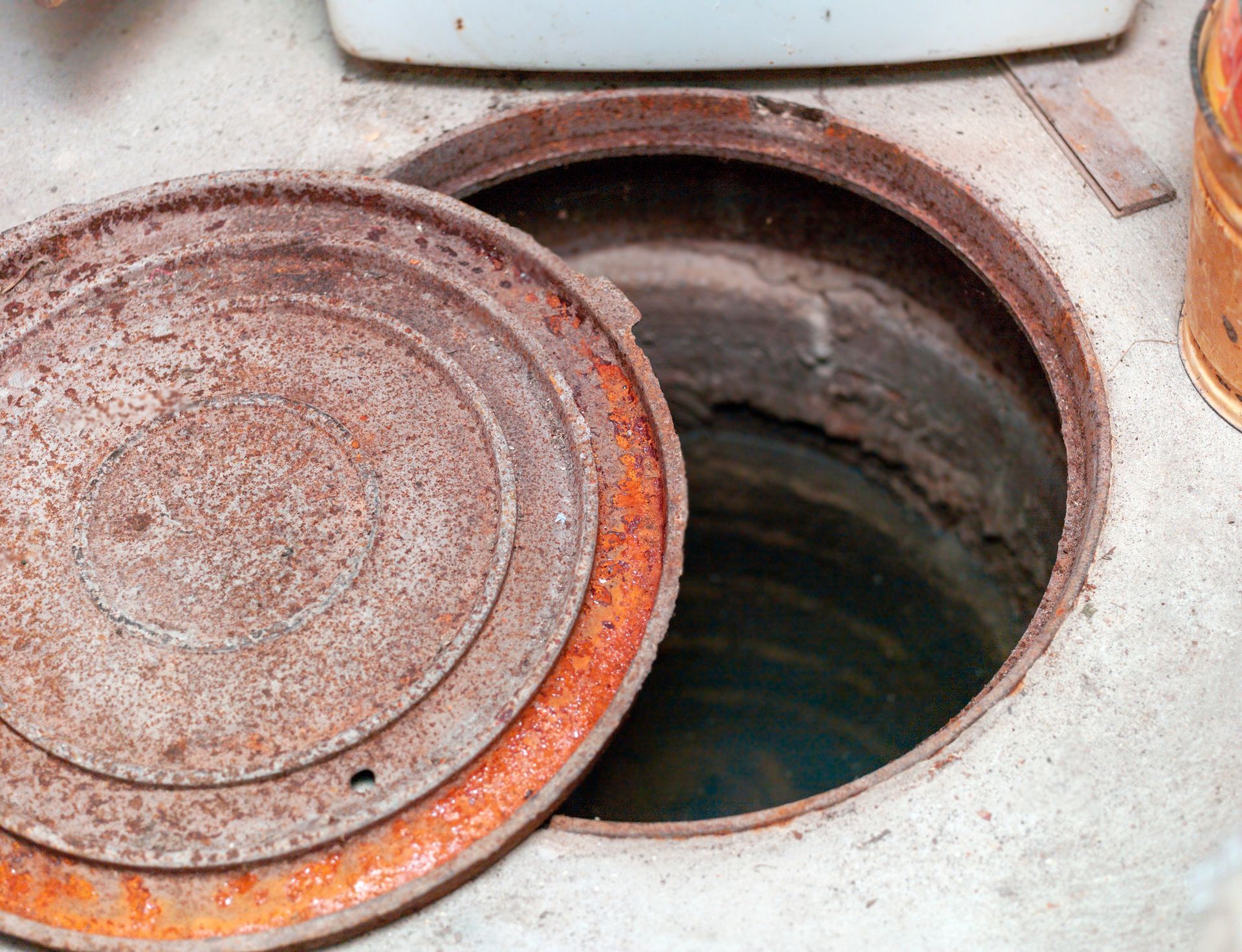 Open rusty manhole cover revealing a dark, cylindrical hole in a concrete floor.