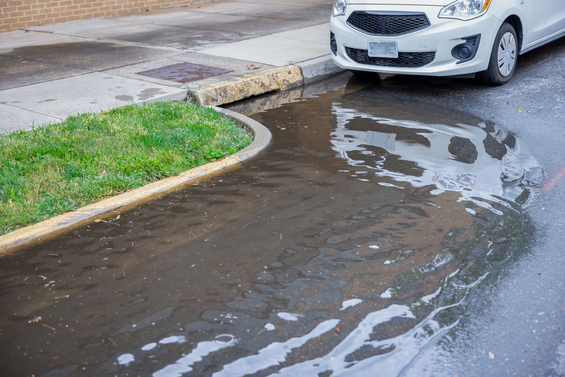 Puddle on asphalt near a curb and grass. A white car is parked nearby.