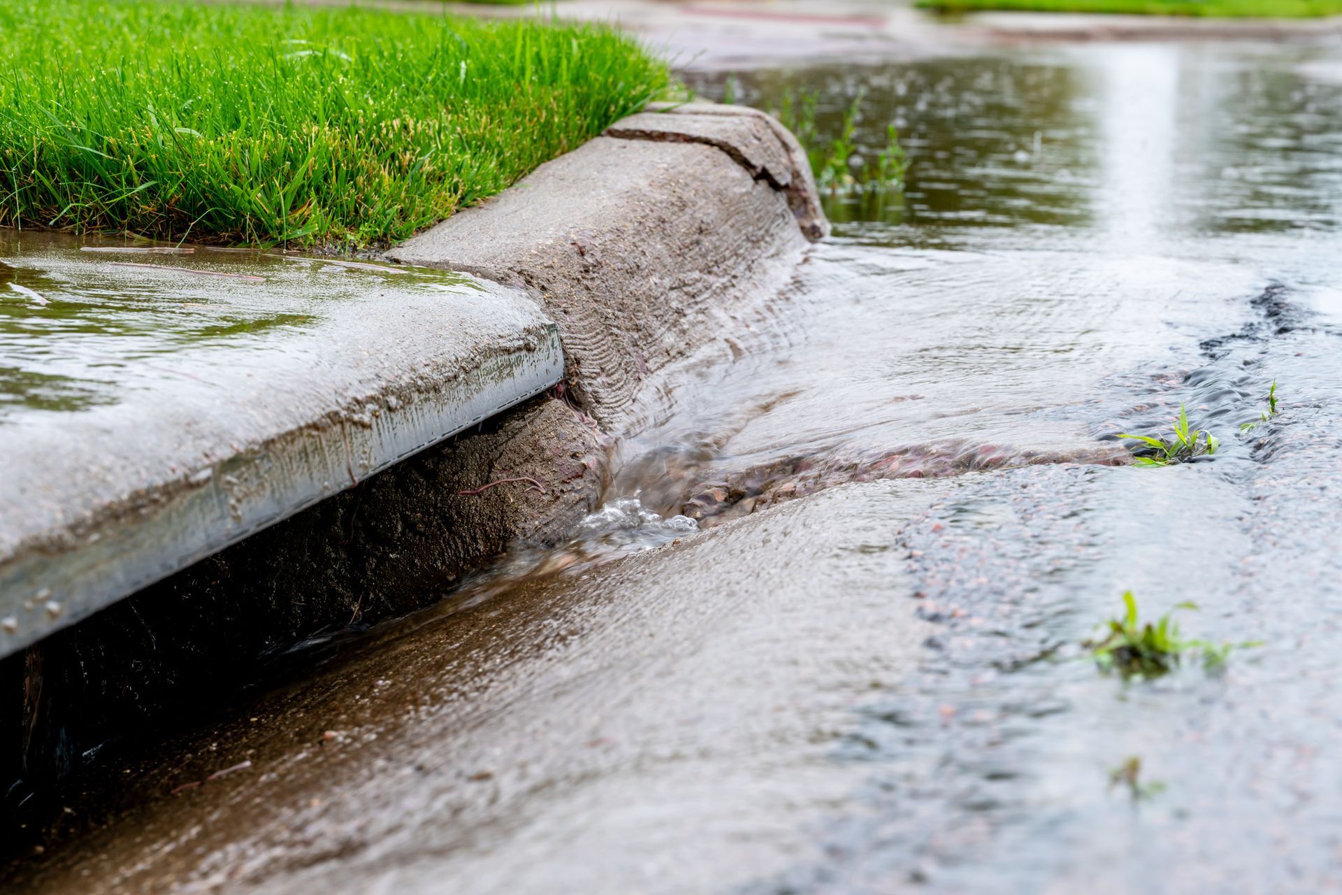 Water overflowing from a curb onto a flooded street, beside green grass.