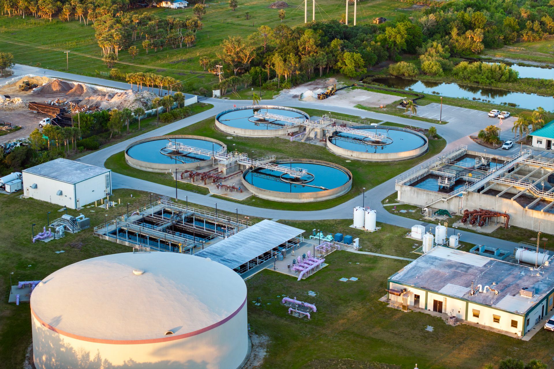 Aerial view of a wastewater treatment plant with several circular tanks and buildings, located near green vegetation and water.