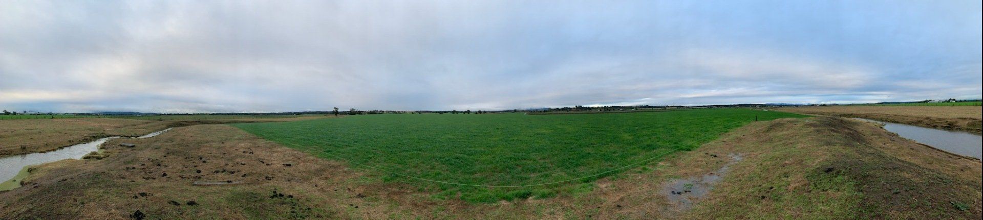 A vast green field surrounded by brown dirt and small waterways under a cloudy sky.