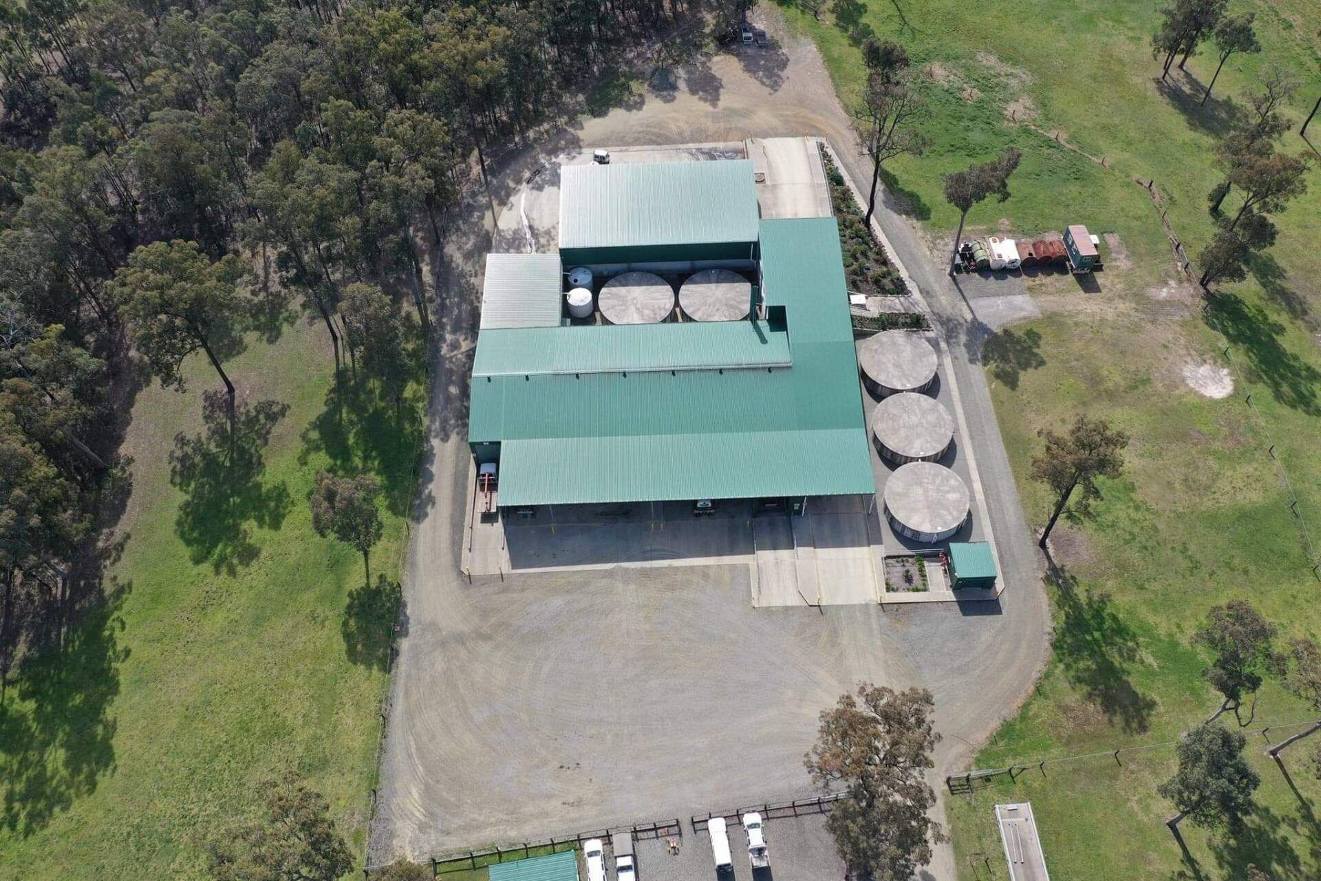 Aerial view of a green-roofed building and silos on a gravel lot, surrounded by trees and grass.