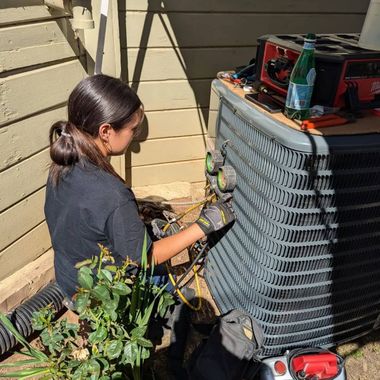 Woman in work clothes repairs AC unit outside, using tools and gauges.