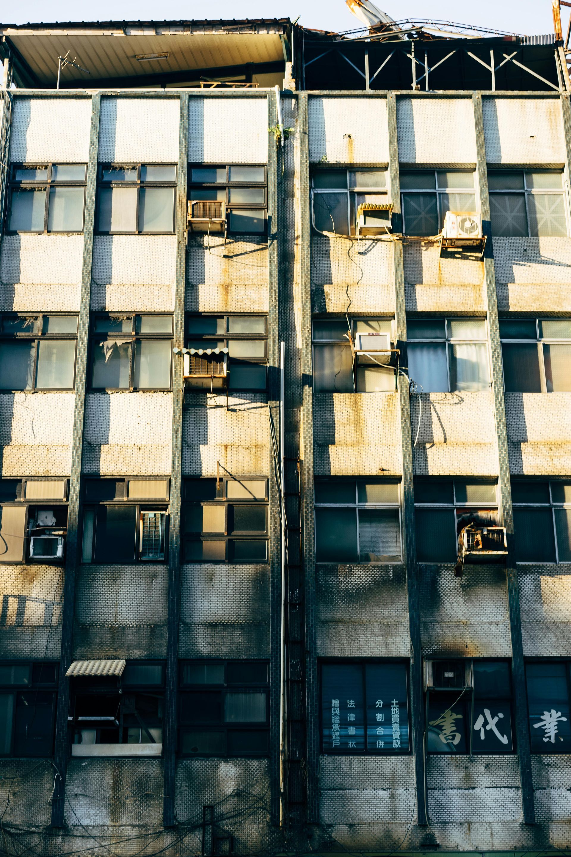 A weathered, multi-story concrete building with numerous square windows, many with air conditioners, in a city setting.