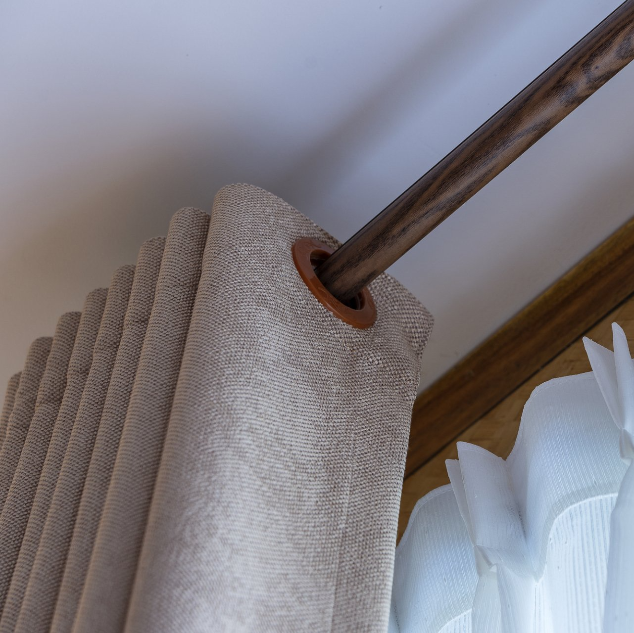 Close up of eyelet curtains pulled to side of wooden curtain rod, over white sheer curtain.