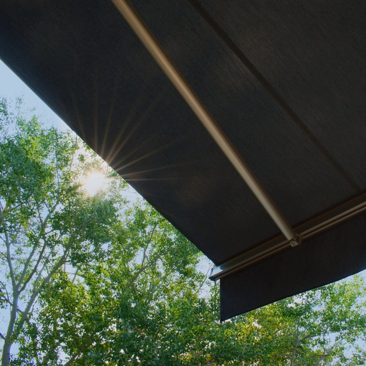 Close up of corner of black outdoor awning, with sun shining through trees in the background.