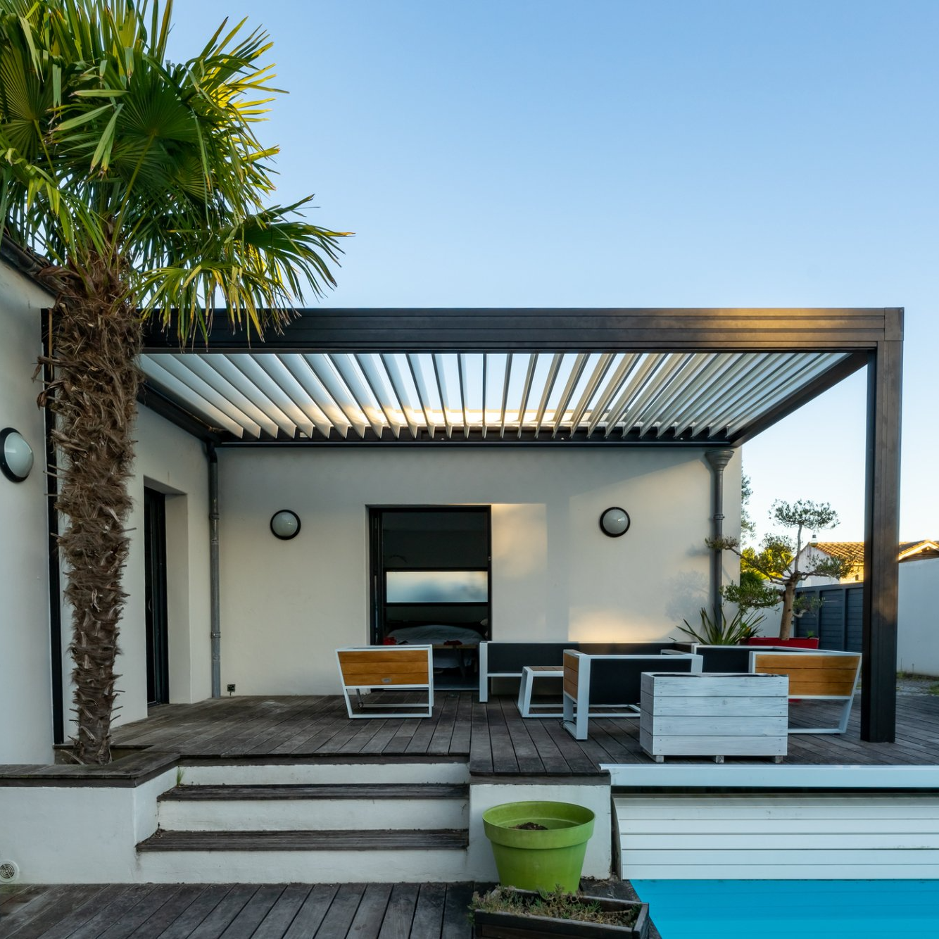 Fixed outdoor awning over decked house patio overlooking pool, framed by palm tree.