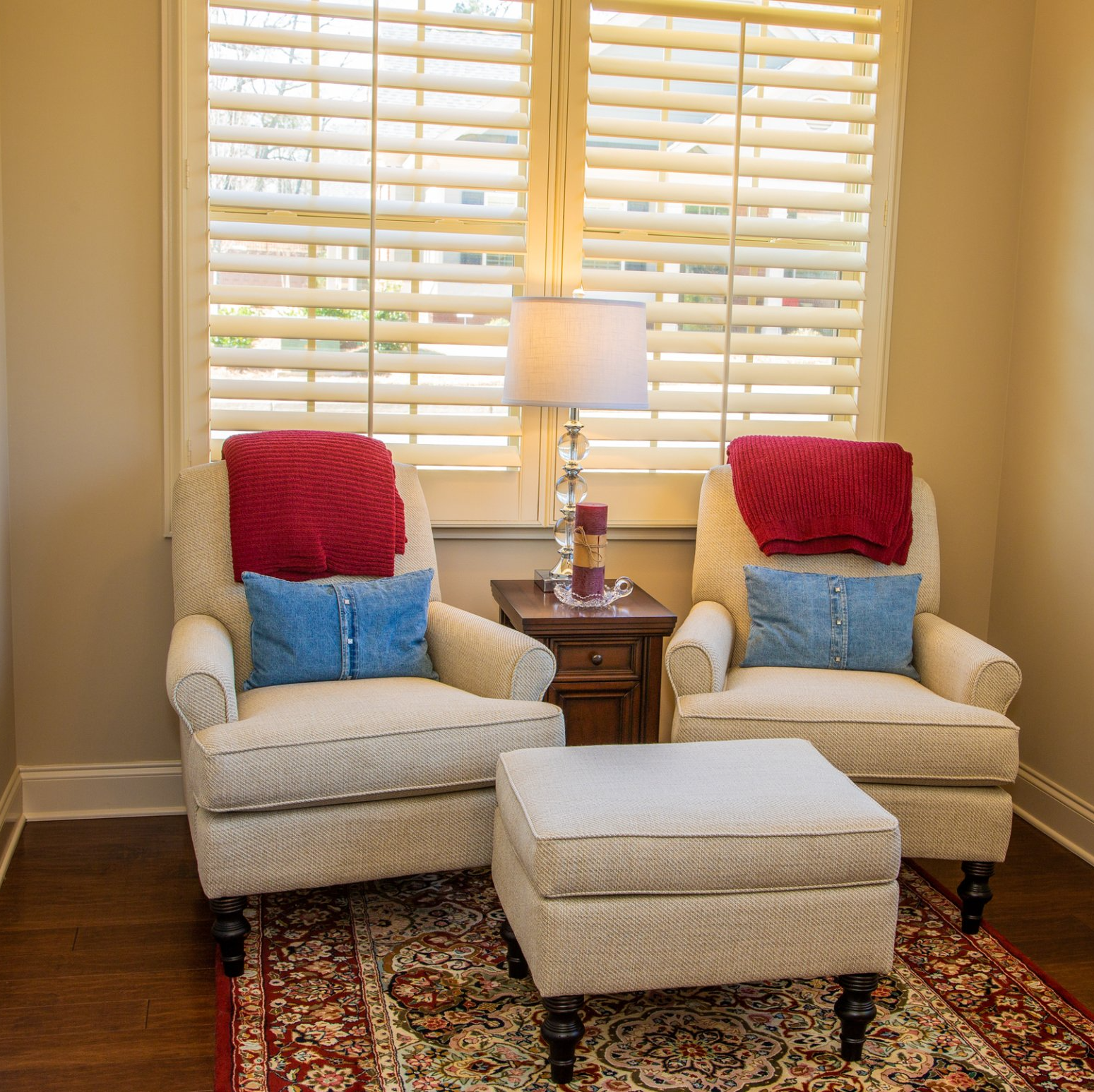 Lounge with footrest and two formal chairs on rug. Chairs have bright red and blue cushions. Open Plantation shutters in background.