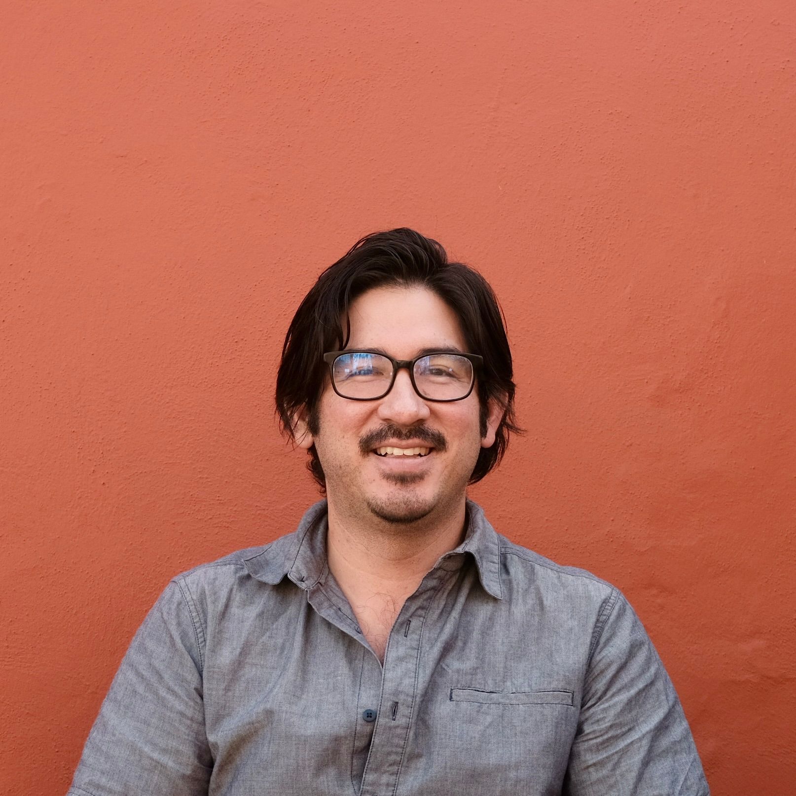 A man in a grey collared shirt with black glasses and a gentle smile