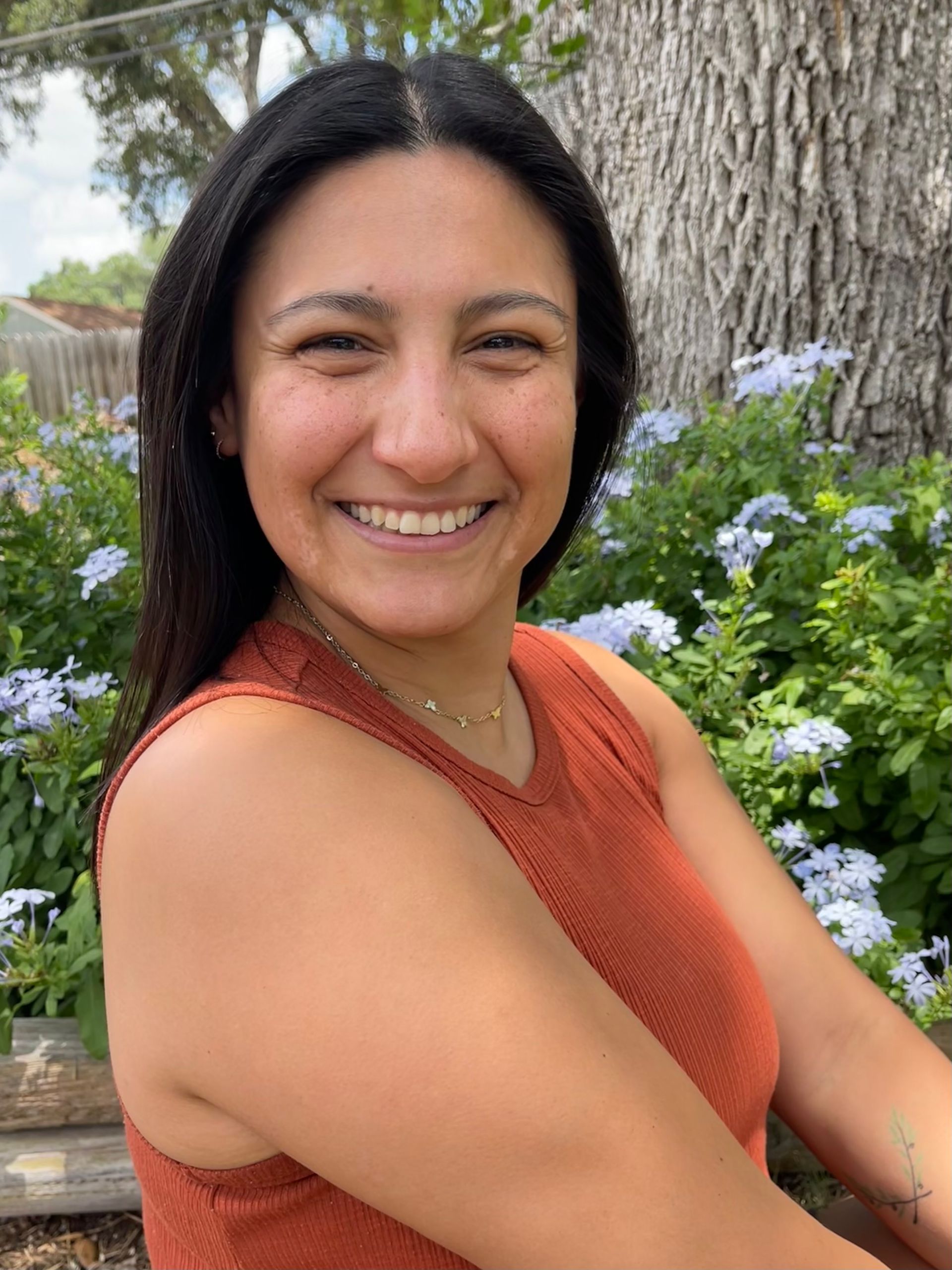 A woman in a red tank top is smiling in front of a bush of flowers.