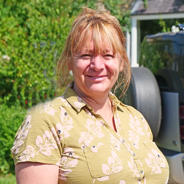 Woman with blonde hair and a patterned olive green shirt smiling outdoors, near greenery.