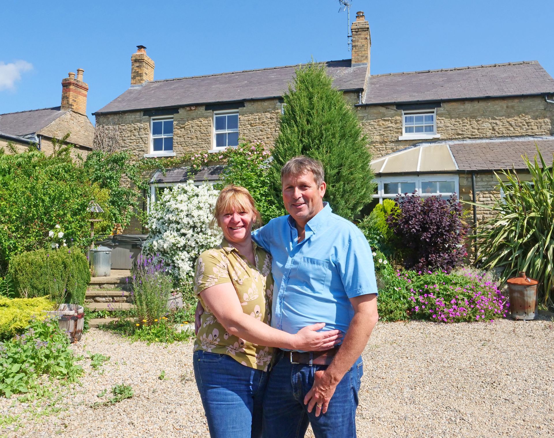 A smiling couple embraces in front of a stone house with a garden. They are outdoors on a sunny day.