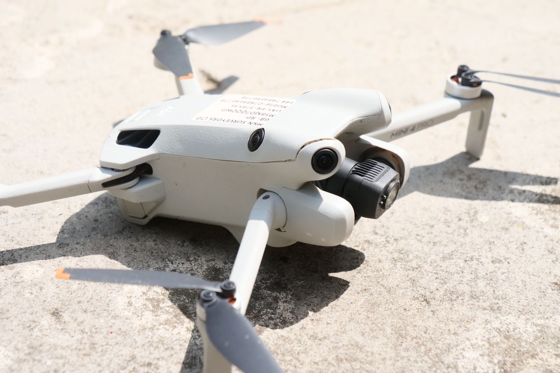 A white drone with its blades extended rests on a light-colored surface, viewed at eye level. The camera is black.