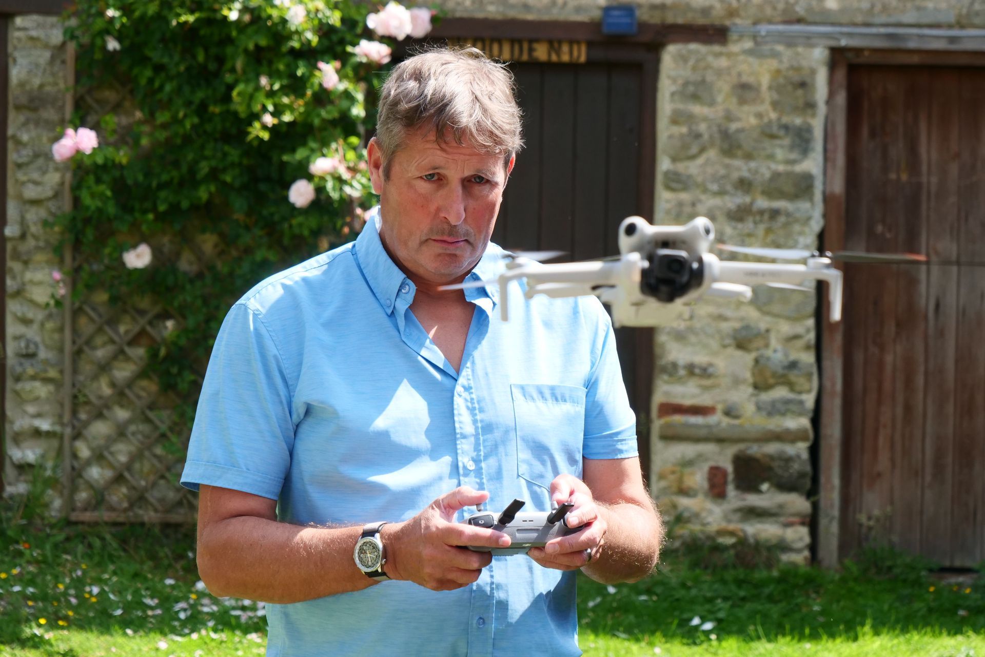 Man in blue shirt operating a drone in a garden, stone building and rose bushes in the background.