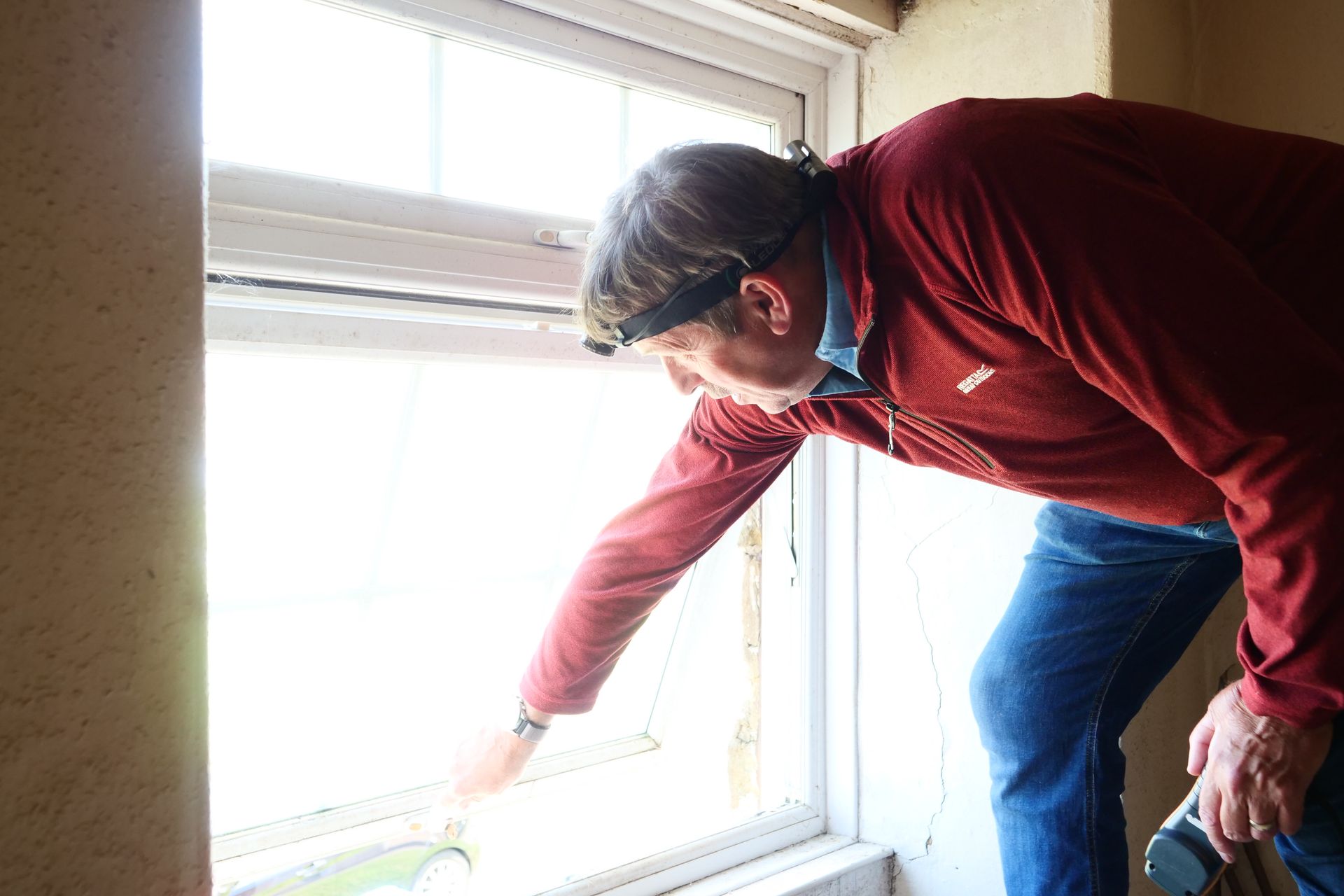Man inspecting a window, reaching out with his hand. He's wearing a headlamp, red jacket, and blue jeans, inside a building.
