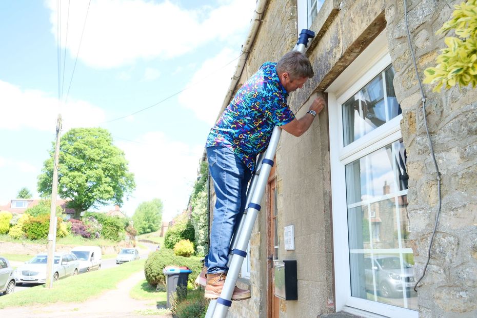 Man on a ladder installing something around a window on the side of a stone house. He's wearing a colorful shirt and pants.