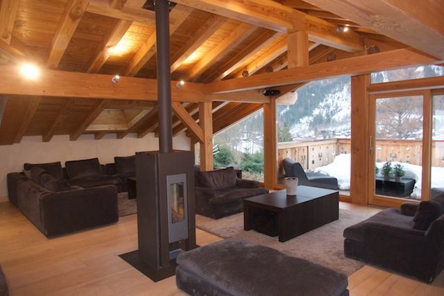 A cozy, wooden-beamed living room with dark brown furniture, a modern fireplace, and a snowy view outside a glass door.