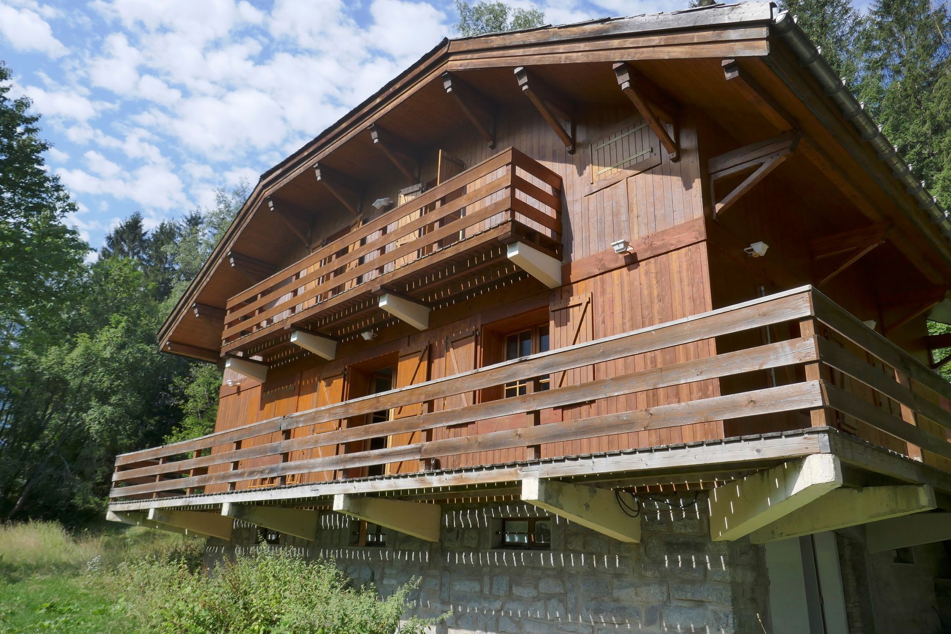 Wooden chalet with balconies, set in a grassy area with trees and a cloudy sky.
